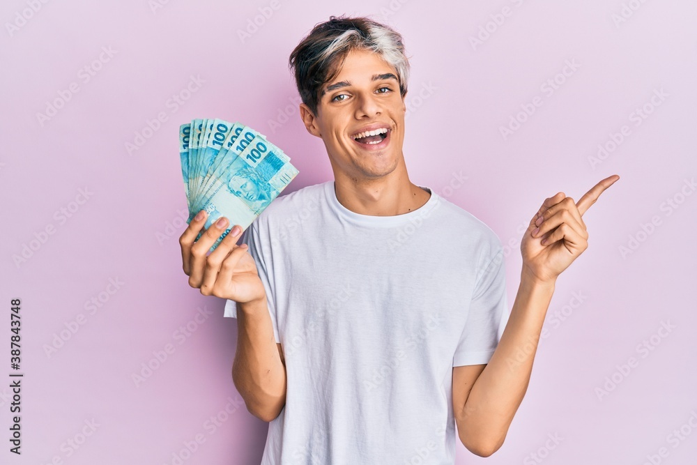 Young hispanic man holding 100 brazilian real banknotes smiling happy pointing with hand and finger to the side Young hispanic man holding 100 brazilian real banknotes smiling happy pointing with hand and finger to the side