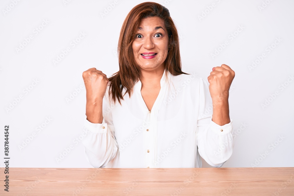 Middle age brunette hispanic business woman wearing casual white shirt sitting on the table celebrating surprised and amazed for success with arms raised and open eyes. winner concept. Middle age brunette hispanic business woman wearing casual white shirt sitting on the table celebrating surprised and amazed for success with arms raised and open eyes. winner concept.