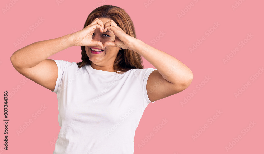 Middle age latin woman wearing casual white tshirt doing heart shape with hand and fingers smiling looking through sign