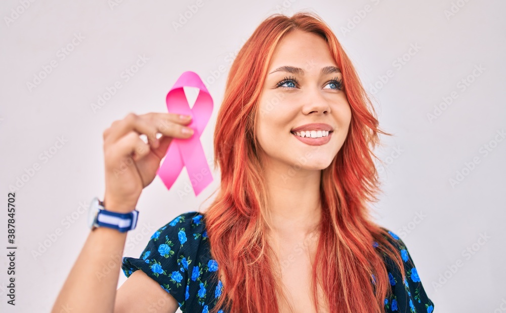 Young redhead girl smiling happy holding breast cancer pink ribbon walking at the city. Young redhead girl smiling happy holding breast cancer pink ribbon walking at the city.