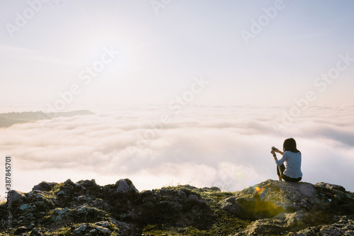 Brunette woman sits on the cliff holds smartphone and takes a photo of the sunrise above the clouds. Concept of travel joy and memories.