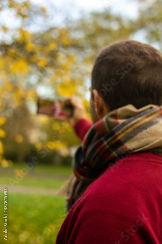 Photo of a young and attractive man in the park during autumn season taking a selfie with his phone. He is wearing a scarf and is smiling. Surrounded by trees with yellow, green and orange colours.