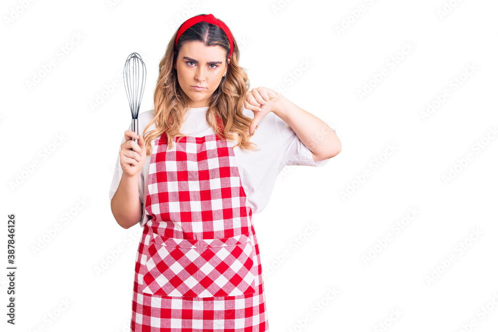 Young caucasian woman wearing apron holding whisk with angry face, negative sign showing dislike with thumbs down, rejection concept Young caucasian woman wearing apron holding whisk with angry face, negative sign showing dislike with thumbs down, rejection concept