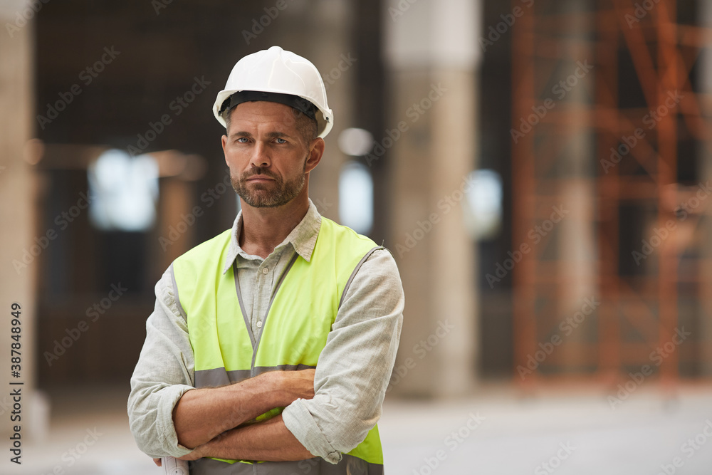 Waist up portrait of mature construction worker looking at camera while standing with arms crossed at construction site, copy space - obrazy, fototapety, plakaty 