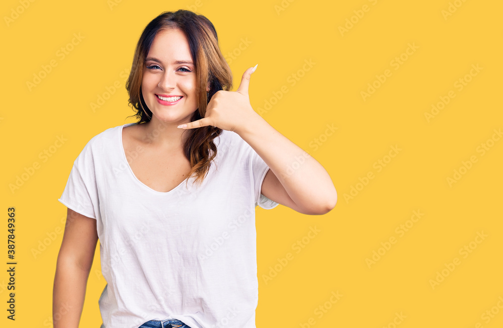 Young beautiful caucasian woman wearing casual white tshirt smiling doing phone gesture with hand and fingers like talking on the telephone. communicating concepts. Young beautiful caucasian woman wearing casual white tshirt smiling doing phone gesture with hand and fingers like talking on the telephone. communicating concepts.