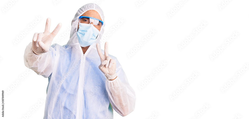 Young hispanic man wearing doctor protection coronavirus uniform and medical mask smiling looking to the camera showing fingers doing victory sign. number two.