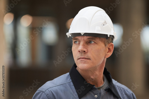 Close up portrait of mature worker wearing hardhat and looking away while standing at construction site or in industrial workshop, copy space
