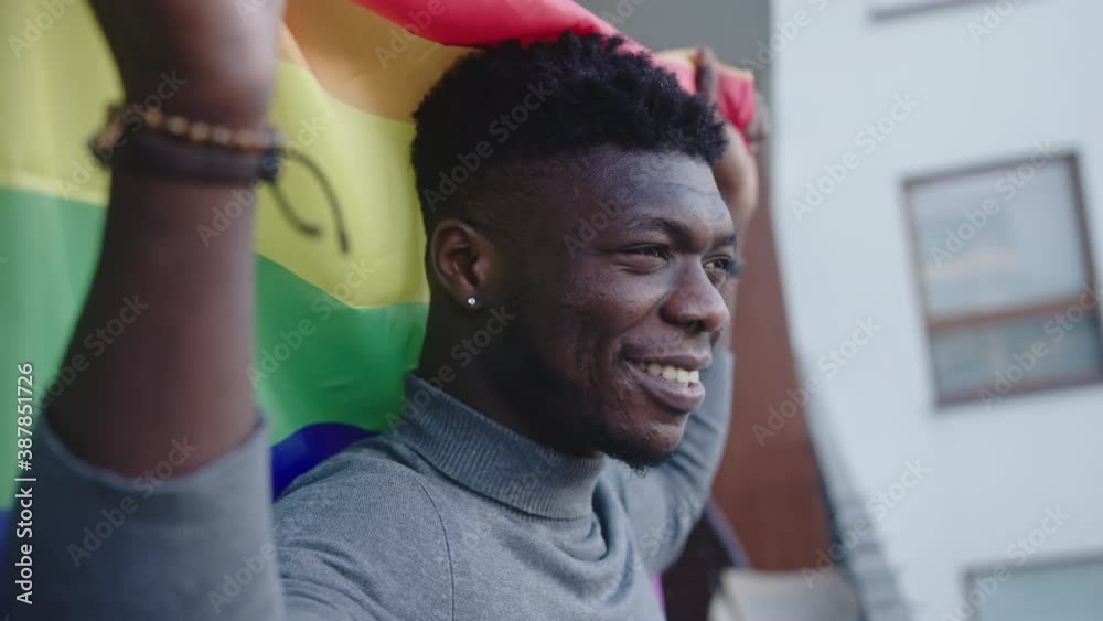 Young handsome homosexual black man holding rainbow flag on the balcony. High quality 4k footage