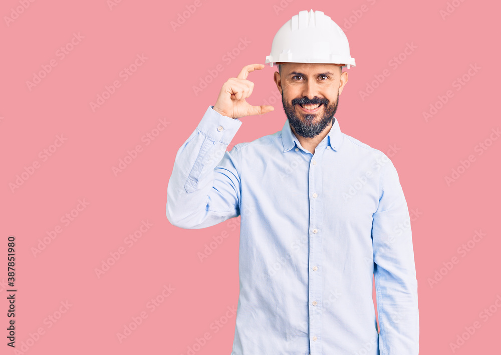 Young handsome man wearing architect hardhat smiling and confident gesturing with hand doing small size sign with fingers looking and the camera. measure concept. Young handsome man wearing architect hardhat smiling and confident gesturing with hand doing small size sign with fingers looking and the camera. measure concept.