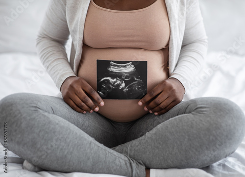 Unrecognizable black pregnant lady demonstrating her baby sonography photo, sitting on bed