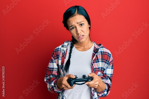 Beautiful hispanic woman playing video game holding controller in shock face, looking skeptical and sarcastic, surprised with open mouth