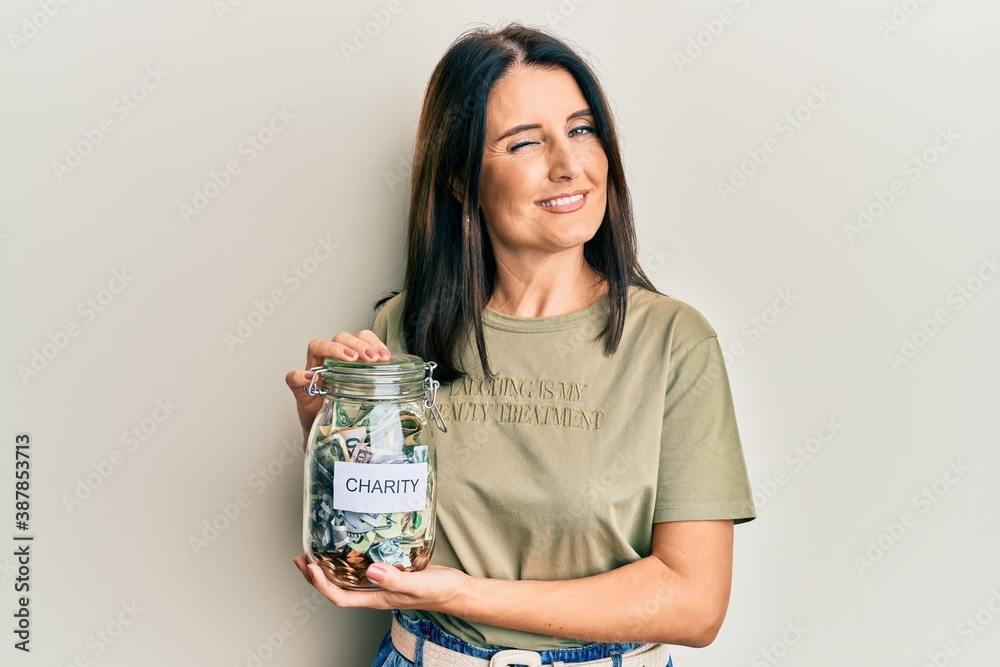Middle age brunette woman holding charity jar with money winking looking at the camera with sexy expression, cheerful and happy face. Middle age brunette woman holding charity jar with money winking looking at the camera with sexy expression, cheerful and happy face.