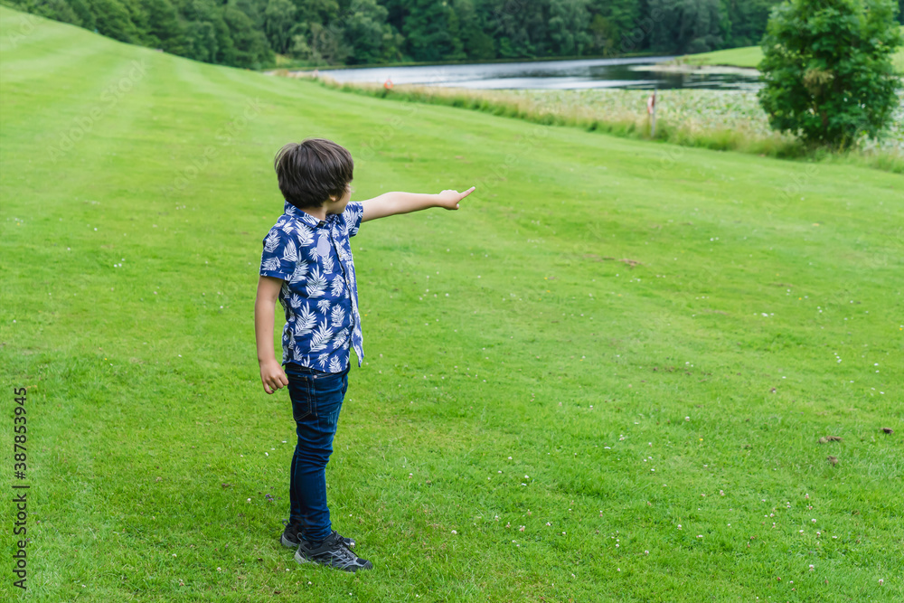 Sideview of Portrait kid standing on green fields pointing finger out. Happy boy playing outdoor in the summer field, Active child standing alone relaxing outside in spring time...
