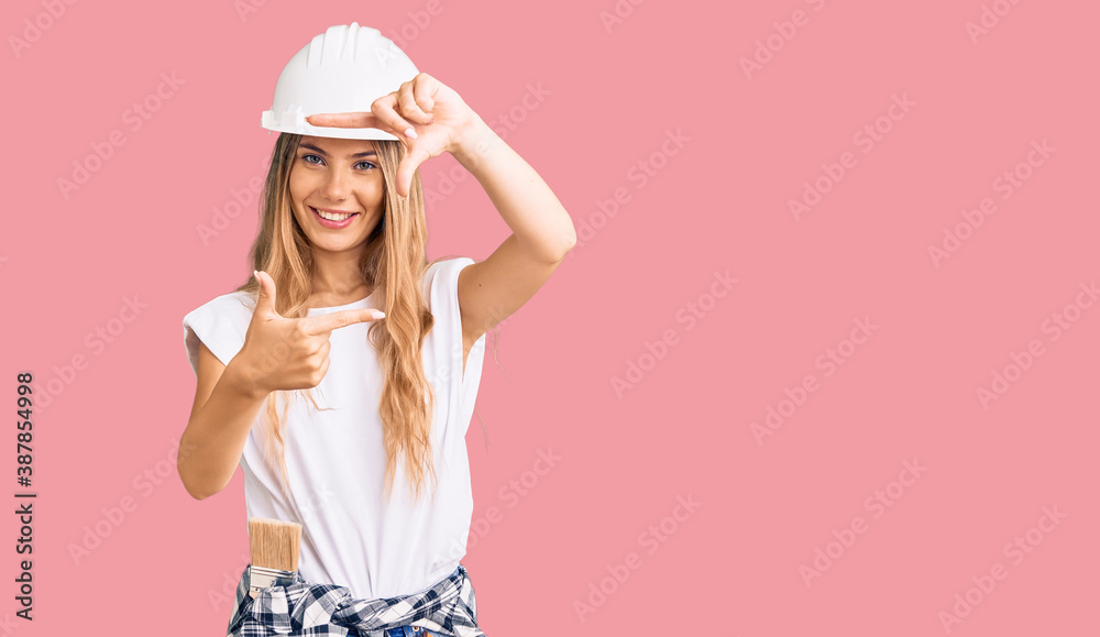 Beautiful caucasian woman with blonde hair wearing hardhat and painter clothes smiling making frame with hands and fingers with happy face. creativity and photography concept. Beautiful caucasian woman with blonde hair wearing hardhat and painter clothes smiling making frame with hands and fingers with happy face. creativity and photography concept.
