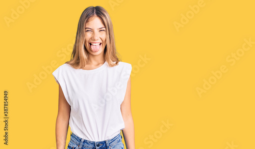 Beautiful caucasian woman with blonde hair wearing casual white tshirt sticking tongue out happy with funny expression. emotion concept.