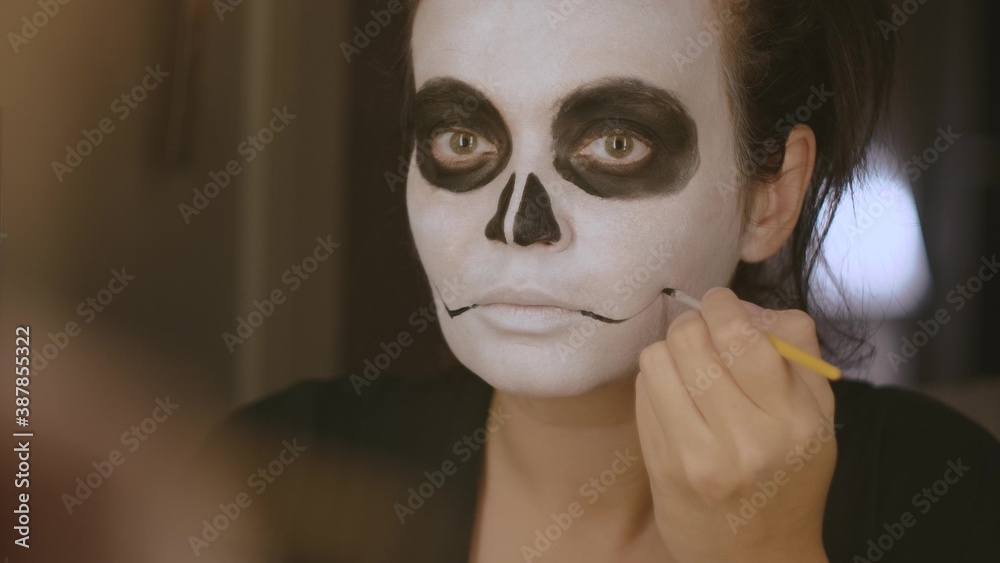 A brunette girl with a white tone on her face, a black nose and circles on her eyes with a thin brush draws arrows at the mouth. Close-up. Halloween makeup. October holiday. A brunette girl with a white tone on her face, a black nose and circles on her eyes with a thin brush draws arrows at the mouth. Close-up. Halloween makeup. October holiday.