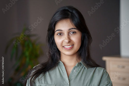 Smiling young adult indian woman looking at camera at home or in office. Pretty lady standing in India house apartment, teacher, real estate agent, housewife close up face front headshot portrait.