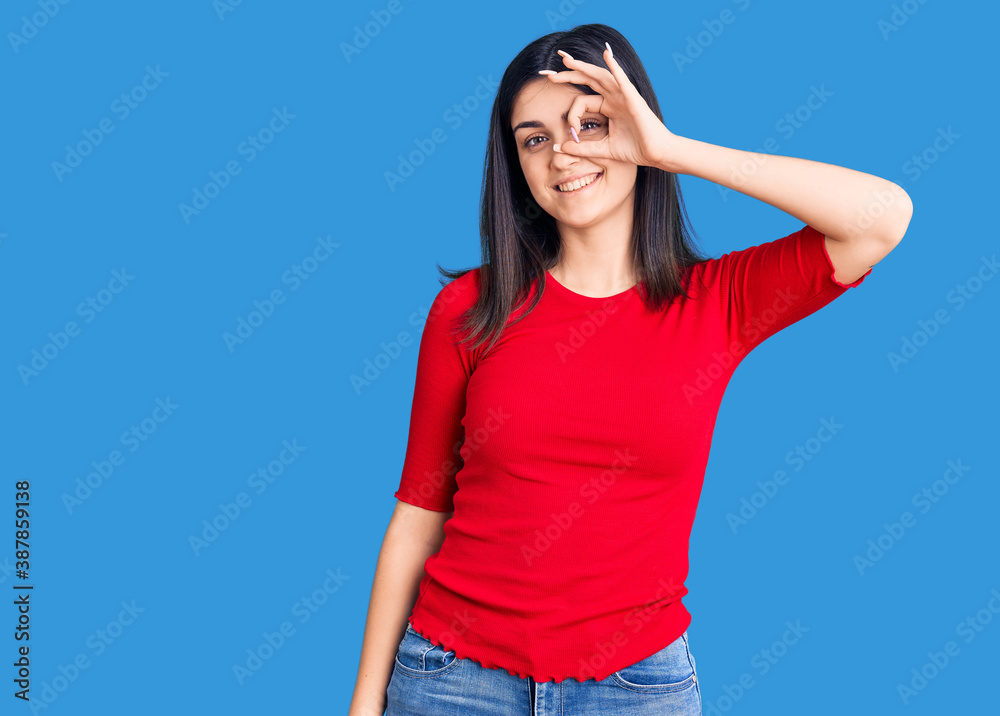 Young beautiful girl wearing casual t shirt smiling happy doing ok sign with hand on eye looking through fingers Young beautiful girl wearing casual t shirt smiling happy doing ok sign with hand on eye looking through fingers