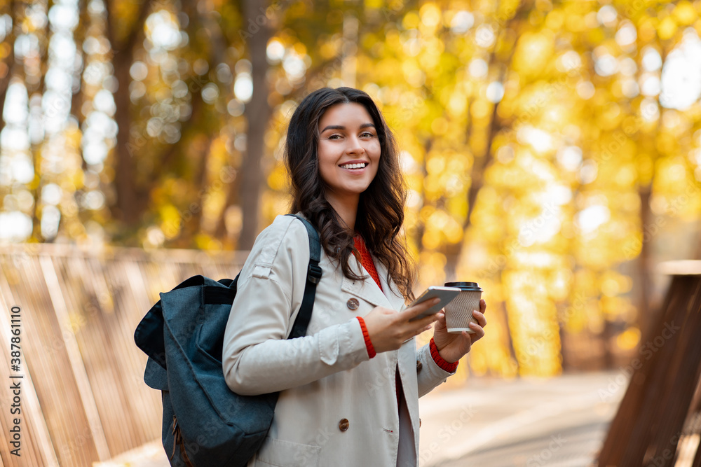 Happy millennial woman with smartphone, takeaway coffee and backpack on walk at autumn park