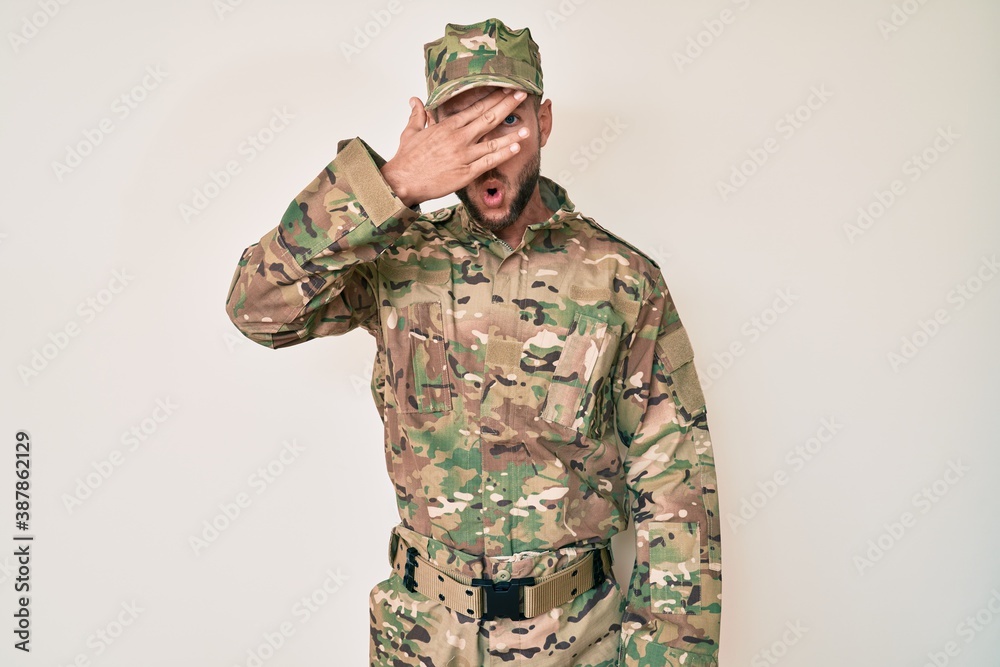 Young caucasian man wearing camouflage army uniform peeking in shock covering face and eyes with hand, looking through fingers with embarrassed expression. Young caucasian man wearing camouflage army uniform peeking in shock covering face and eyes with hand, looking through fingers with embarrassed expression.