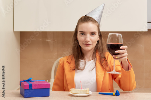 A young woman says a toast in a festive hat and with a glass of wine in her hands. On the table is a birthday cake with a candle.