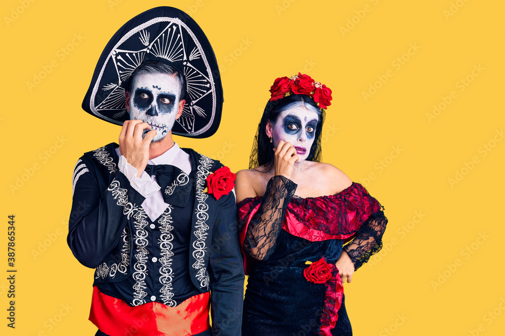 Young couple wearing mexican day of the dead costume over background looking stressed and nervous with hands on mouth biting nails. anxiety problem.