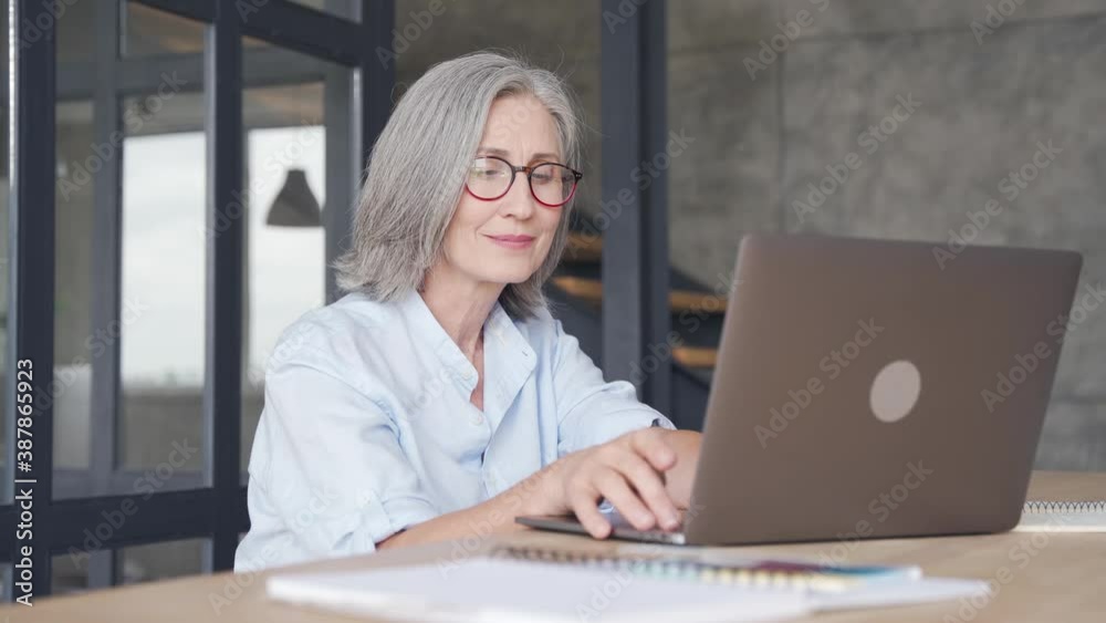 Smiling professional mature business woman wears glasses using laptop computer sits at workplace desk. Happy senior older employee 60s businesswoman executive working typing on pc at home from office.
