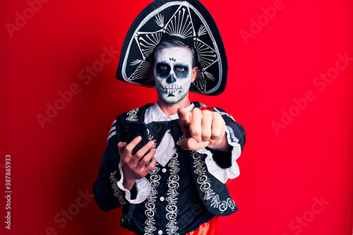 Young man wearing mexican day of the dead costume using smartphone pointing with finger to the camera and to you, confident gesture looking serious