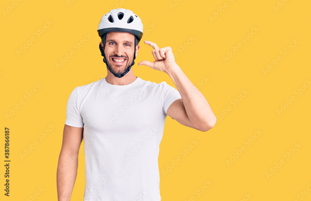 Young handsome man wearing bike helmet smiling and confident gesturing with hand doing small size sign with fingers looking and the camera. measure concept.