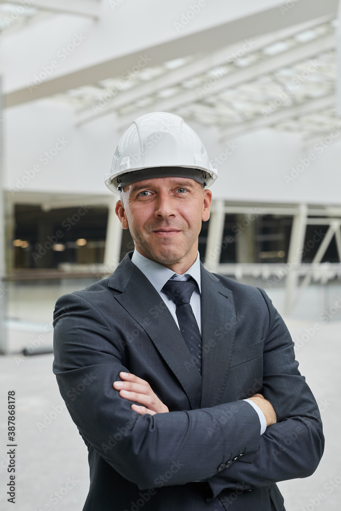 Vertical waist up portrait of smiling mature businessman wearing hardhat and looking at camera while standing with arms crossed at construction site Vertical waist up portrait of smiling mature businessman wearing hardhat and looking at camera while standing with arms crossed at construction site