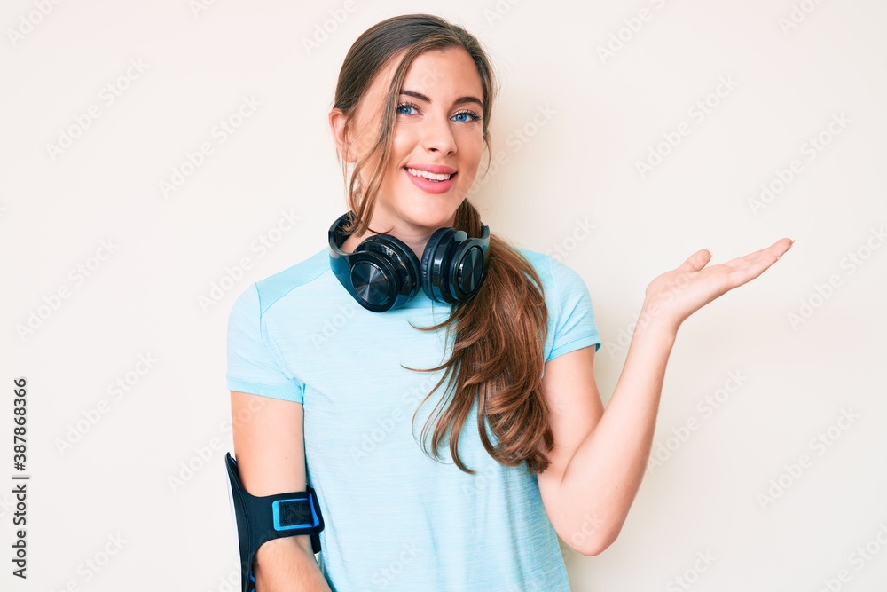 Beautiful young caucasian woman wearing gym clothes and using headphones smiling cheerful presenting and pointing with palm of hand looking at the camera. Beautiful young caucasian woman wearing gym clothes and using headphones smiling cheerful presenting and pointing with palm of hand looking at the camera.