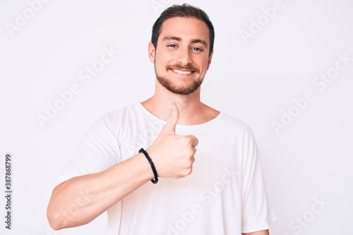 Young handsome man wearing casual white tshirt doing happy thumbs up gesture with hand. approving expression looking at the camera showing success.