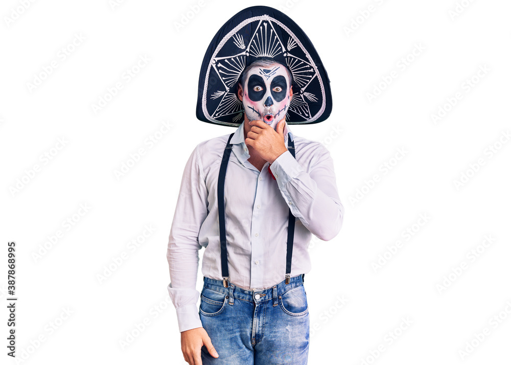 Man wearing day of the dead costume over background looking fascinated with disbelief, surprise and amazed expression with hands on chin Man wearing day of the dead costume over background looking fascinated with disbelief, surprise and amazed expression with hands on chin