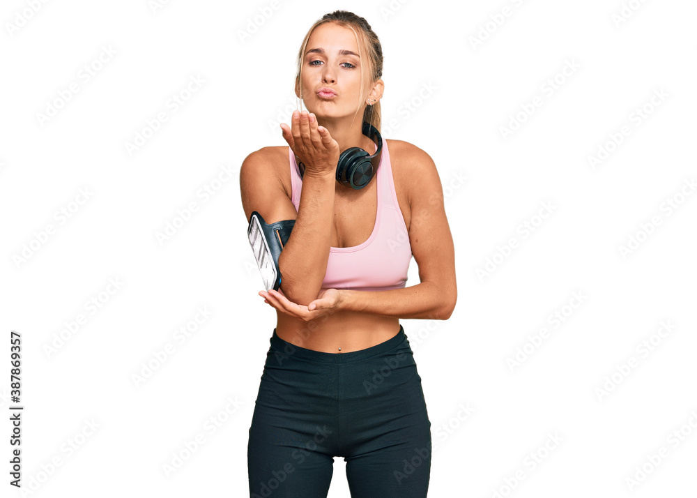 Young blonde woman wearing gym clothes and using headphones looking at the camera blowing a kiss with hand on air being lovely and sexy. love expression. Young blonde woman wearing gym clothes and using headphones looking at the camera blowing a kiss with hand on air being lovely and sexy. love expression.