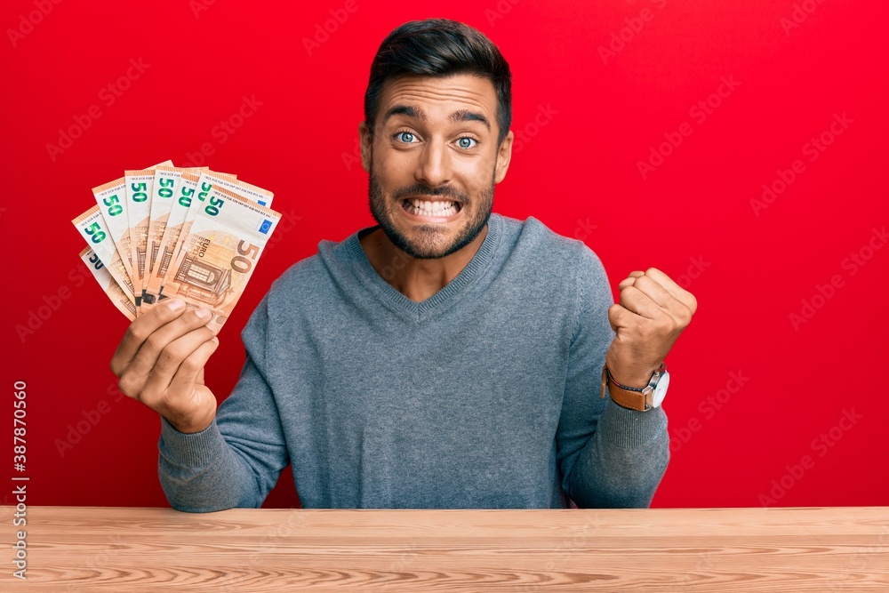 Handsome hispanic man holding bunch of 50 euro banknotes screaming proud, celebrating victory and success very excited with raised arm Handsome hispanic man holding bunch of 50 euro banknotes screaming proud, celebrating victory and success very excited with raised arm