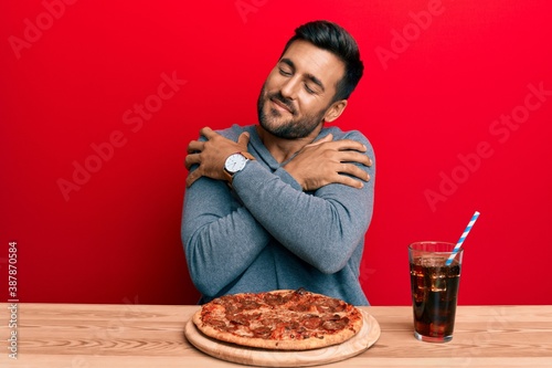 Handsome hispanic man eating tasty pepperoni pizza hugging oneself happy and positive, smiling confident. self love and self care