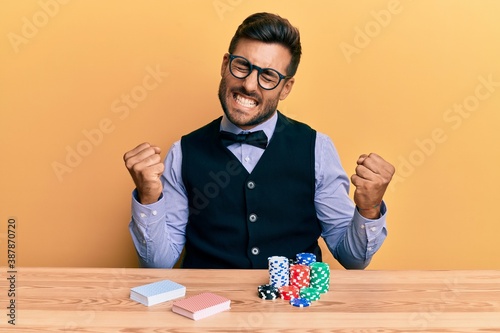 Handsome hispanic croupier man sitting on the table with poker chips and cards very happy and excited doing winner gesture with arms raised, smiling and screaming for success. celebration concept.