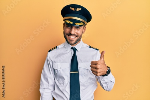 Handsome hispanic man wearing airplane pilot uniform doing happy thumbs up gesture with hand. approving expression looking at the camera showing success. © Krakenimages.com
