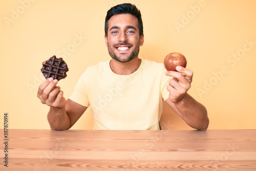 Young hispanic man eating breakfast holding chocolate waflle and apple smiling with a happy and cool smile on face. showing teeth. photo