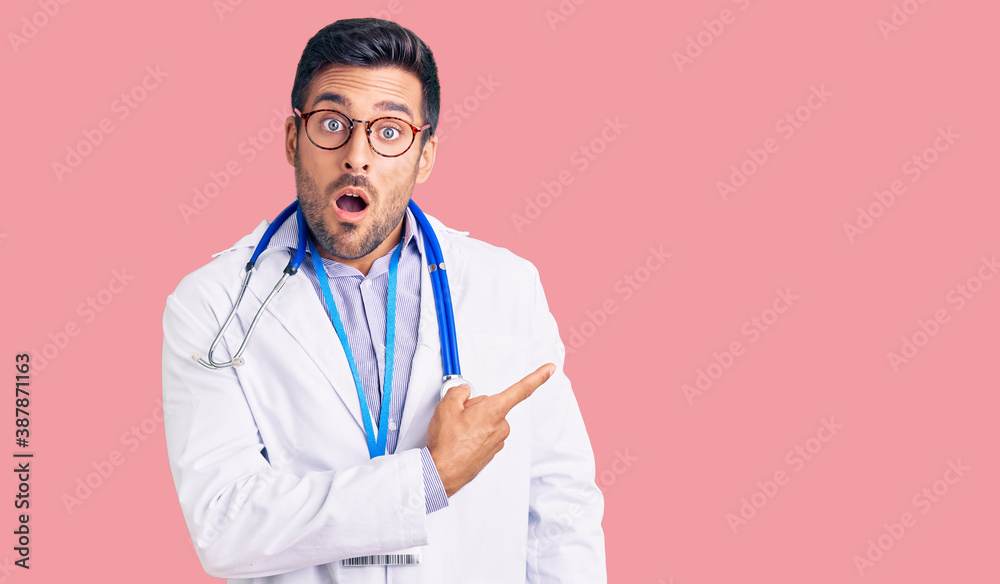 Fotografie Young hispanic man wearing doctor uniform and stethoscope surprised pointing with finger to the side, open mouth amazed expression