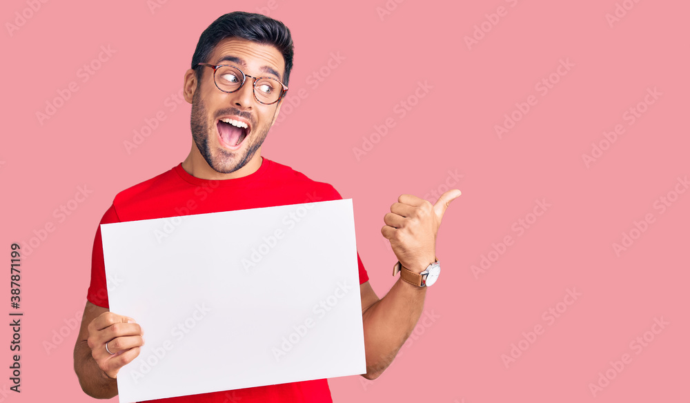 Young hispanic man holding blank empty banner pointing thumb up to the side smiling happy with open mouth Young hispanic man holding blank empty banner pointing thumb up to the side smiling happy with open mouth