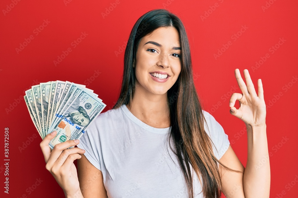 Young brunette woman holding dollars doing ok sign with fingers, smiling friendly gesturing excellent symbol Young brunette woman holding dollars doing ok sign with fingers, smiling friendly gesturing excellent symbol