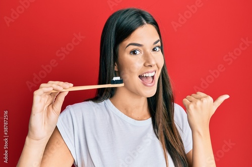 Young brunette woman holding toothbrush with toothpaste pointing thumb up to the side smiling happy with open mouth