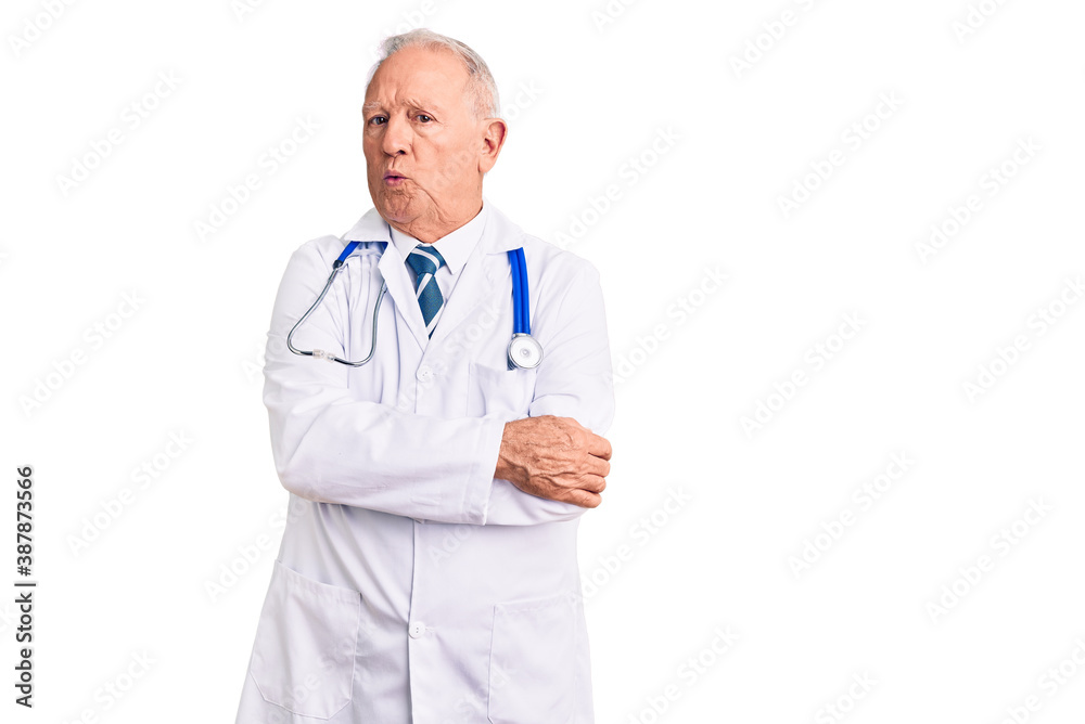 Senior handsome grey-haired man wearing doctor coat and stethoscope shaking and freezing for winter cold with sad and shock expression on face