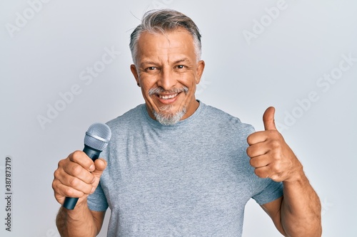 Middle age grey-haired man singing song using microphone smiling happy and positive, thumb up doing excellent and approval sign