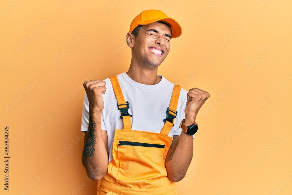 Young handsome african american man wearing handyman uniform over yellow background very happy and excited doing winner gesture with arms raised, smiling and screaming for success. celebration concept Young handsome african american man wearing handyman uniform over yellow background very happy and excited doing winner gesture with arms raised, smiling and screaming for success. celebration concept