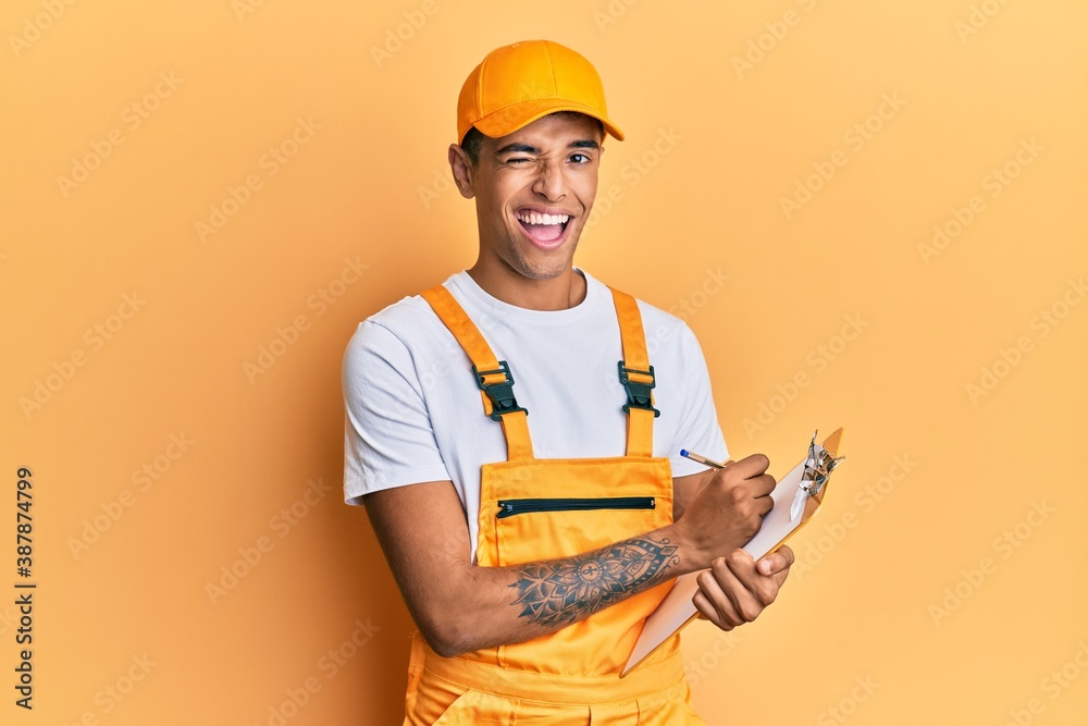 Young handsome african american man wearing courier uniform holding clipboard winking looking at the camera with sexy expression, cheerful and happy face. Young handsome african american man wearing courier uniform holding clipboard winking looking at the camera with sexy expression, cheerful and happy face.