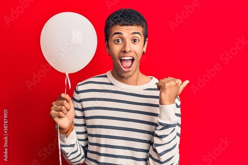 Young african amercian man holding balloon pointing thumb up to the side smiling happy with open mouth