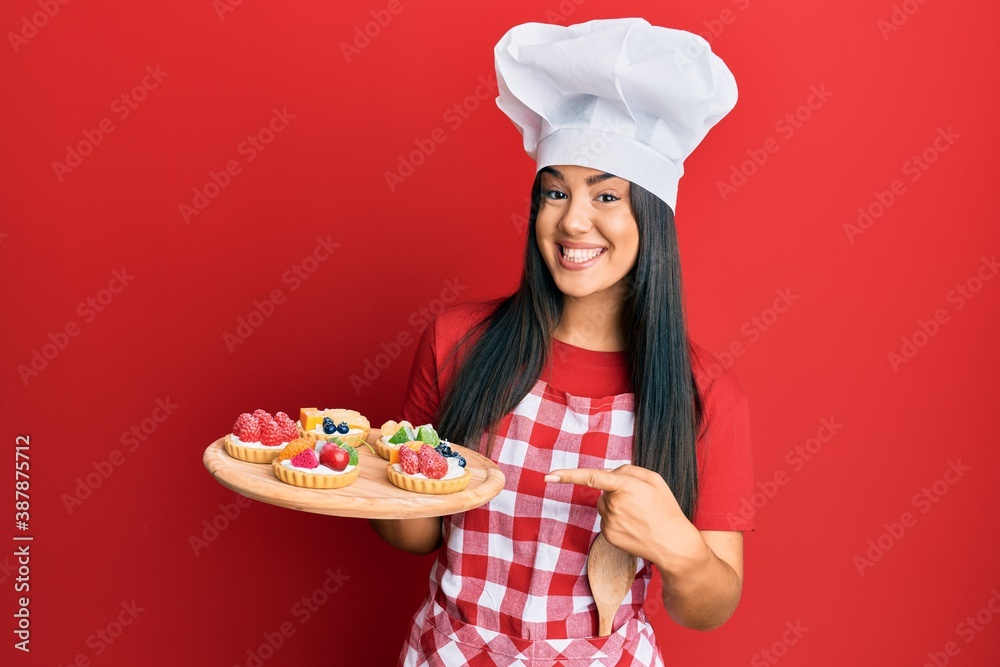Young beautiful hispanic girl wearing baker apron holding cakes smiling happy pointing with hand and finger Young beautiful hispanic girl wearing baker apron holding cakes smiling happy pointing with hand and finger