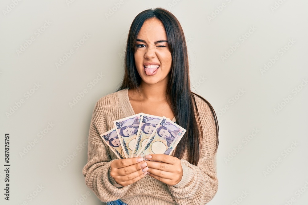 Young beautiful hispanic girl holding japanese yen banknotes sticking tongue out happy with funny expression. Young beautiful hispanic girl holding japanese yen banknotes sticking tongue out happy with funny expression.